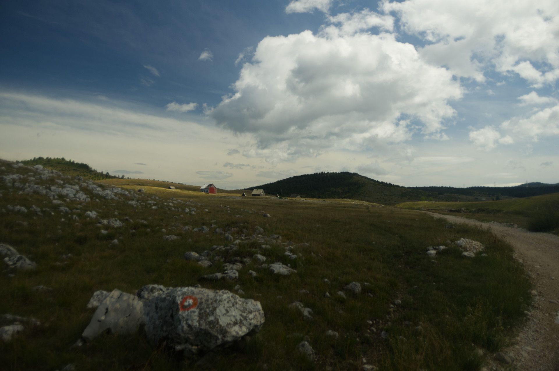A photo of a few houses standing on a hill next to a gravel road with a rock with a trail marker in the foreground